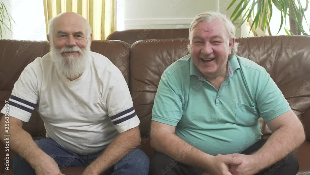 Portrait of two mature senior men sitting on the brown leather sofa. Happy people high five, takes each other hands. Leisure of the pensioners Portrait of two mature senior men sitting on the brown leather sofa. Happy people high five, takes each other hands. Leisure of the pensioners
