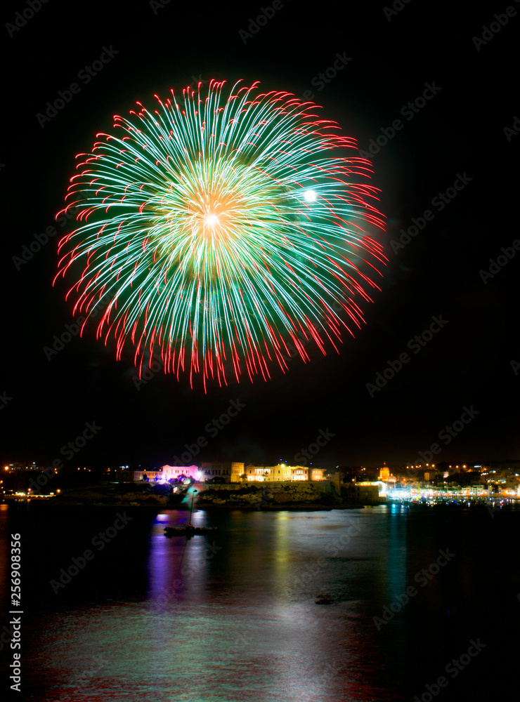 Naklejka premium Colorful fireworks isolated in dark background with the place for text,Malta fireworks festival.Fireworks.Colorful explode with Valletta background, big explosion,reflections on a water in Valleta