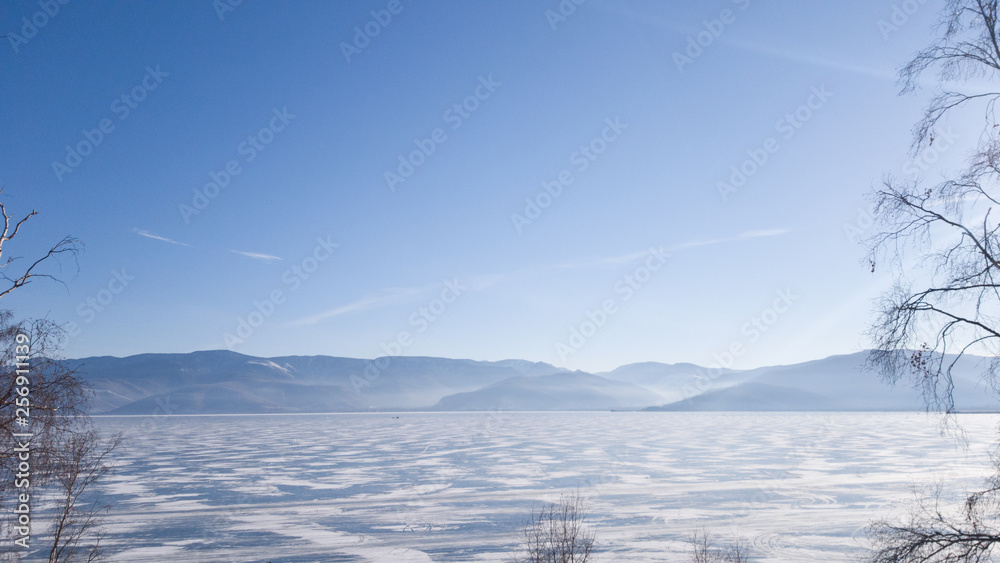 View of the winter lake Baikal: the surface of the lake in the ice, on the other side of the mountain, in the foreground trees. Clear Sunny weather in the winter of Siberia