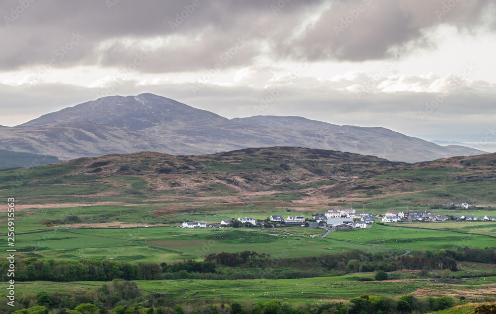 A small village on the island of Jura sits beneath large hills and is surrounded by farmland. Viewed from the island of Islay, Scotland.