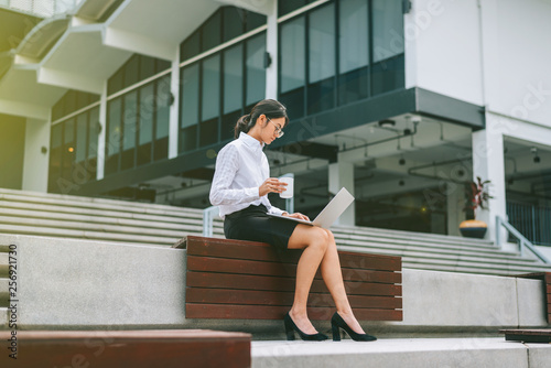 Busy Asian businesswoman using laptop working business- financial while drinking coffee sitting front office building in the morning with sun flare effect- woman officer worker- businesswoman concept
