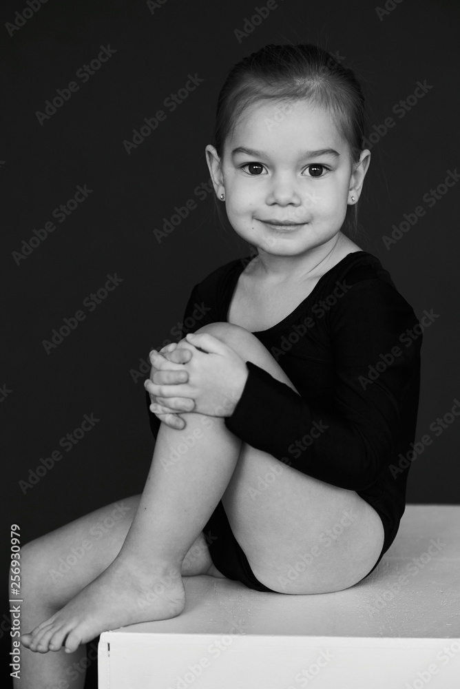A portrait of beautiful little girl dressed in black tights on a white cube against dark background. Adobe Stock