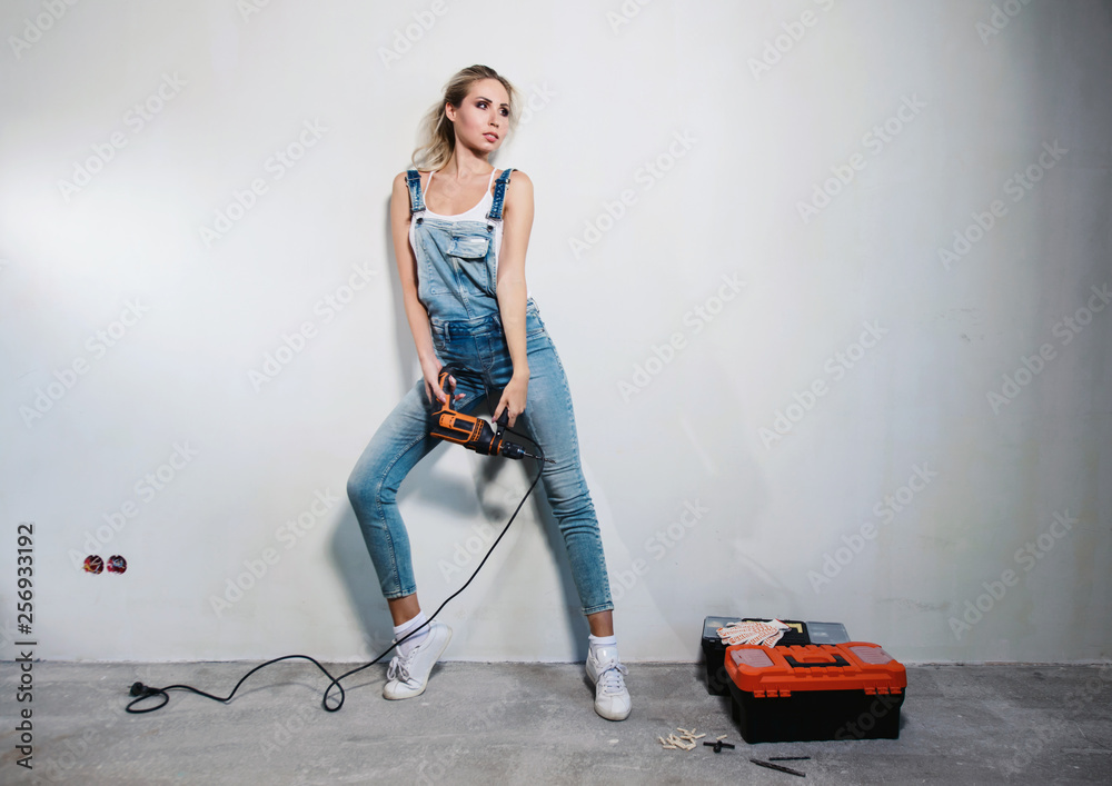 Portrait of young woman worker at construction site with orange drill and  construction tools. Sexy girl using a drill as a gun. Smiling and funny.  Flat or house renovation concept Stock Photo