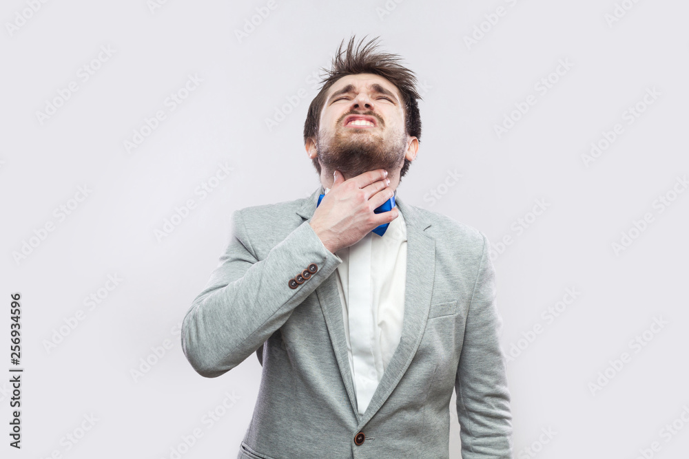 throat pain. Portrait of sick handsome bearded man in casual grey suit and blue bow tie standing and holding his painful throat. indoor studio shot, isolated on light grey background. throat pain. Portrait of sick handsome bearded man in casual grey suit and blue bow tie standing and holding his painful throat. indoor studio shot, isolated on light grey background.