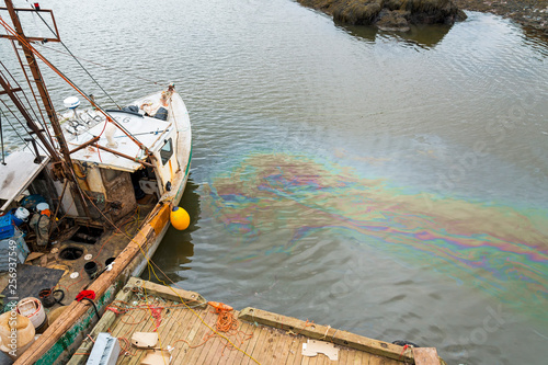 An oil slick in the water, next to an old dilapidated fishing boat. The slick is vibrant and colorful. A dock is visible. 
