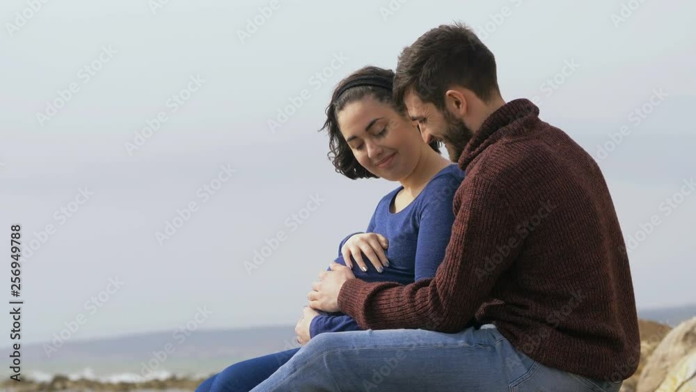 happy young couple sitting on the seashore,hands touching the belly happy young couple sitting on the seashore,hands touching the belly
