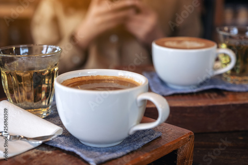 Closeup image of two white cups of hot coffee and a glass of tea on vintage wooden table with a woman in cafe