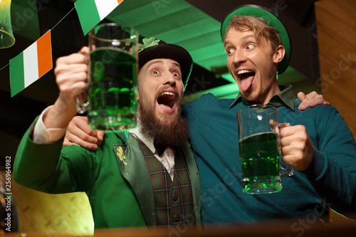 Dark-haired bearded young man in a leprechaun hat and his friend looking drunk