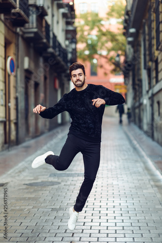 Young bearded man jumping in urban background with open arms wearing casual clothes. 