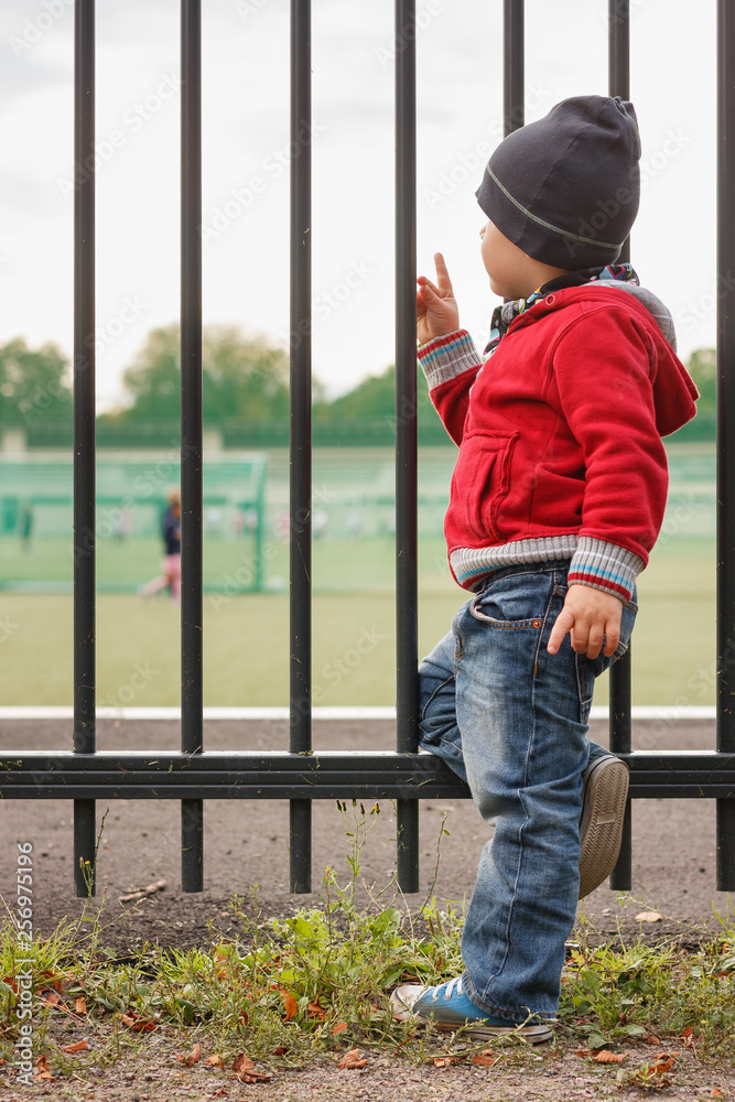 Kid boy stands by the fence and looks into the distance. Metal fence around the stadium or school. The boy looks at the guys playing on the field.