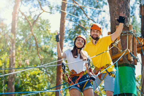 Young woman and man in protective gear are standing on rope bridge hanging on high trees, posing and smiling. Rope park with obstacles and ziplines. Extreme rest and summer activities concept.