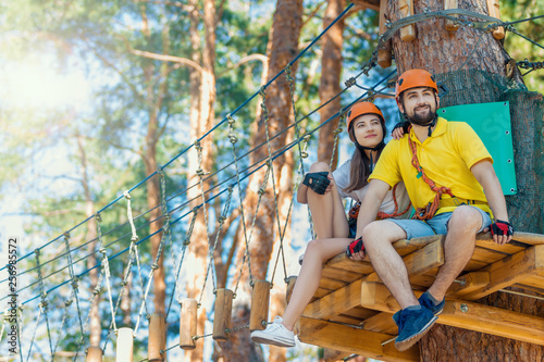 Young woman and man in protective gear are sitting on wooden board on high tree, posing and smiling. Rope adventure park with obstacles and ziplines. Extreme rest and summer activities concept.