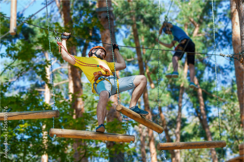 Young male man adult wears protective helmet with action camera having fun in extreme rope park, amusement park. Climbing in rope bridge at green forest. Active healthy lifestyle in spring or summer.