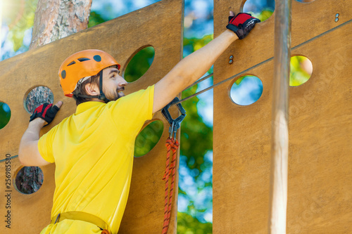 Young male man adult wears protective helmet with action camera having fun in extreme rope park, amusement park. Climbing in wooden wall at green forest. Active healthy lifestyle in spring or summer.