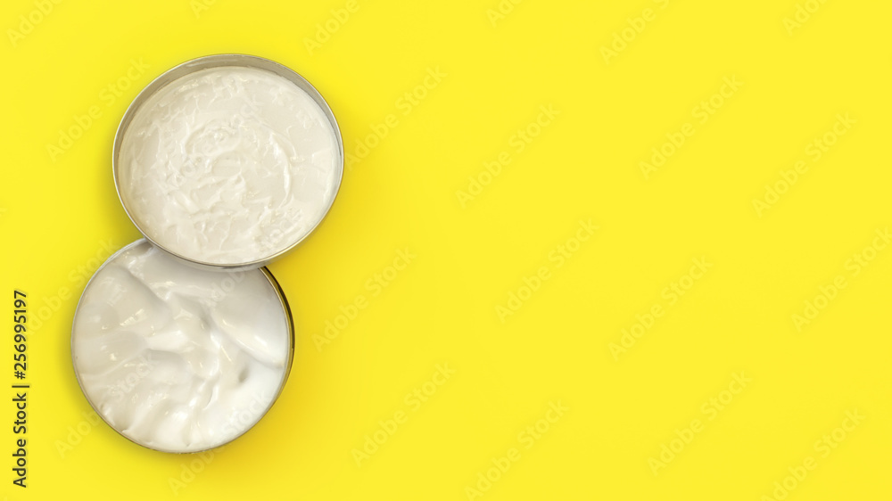 Tabletop view, metal round tin container with white cosmetic cream on yellow board, cap over the jar base, wide banner with text space right,