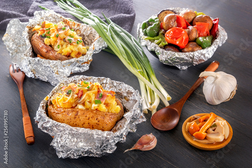 Baked potatoes with bacon, onions and baked vegetables in foil - tomatoes, eggplants, peppers on a gray wooden table.