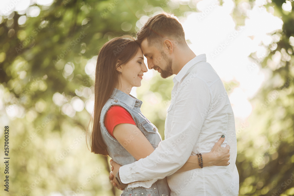 Portrait of young romantic couple in love, smiling and embracing in garden. Sweet lovers in casual clothes, posing touching each other face to face, standing in summer park. Sunset time. Portrait of young romantic couple in love, smiling and embracing in garden. Sweet lovers in casual clothes, posing touching each other face to face, standing in summer park. Sunset time.