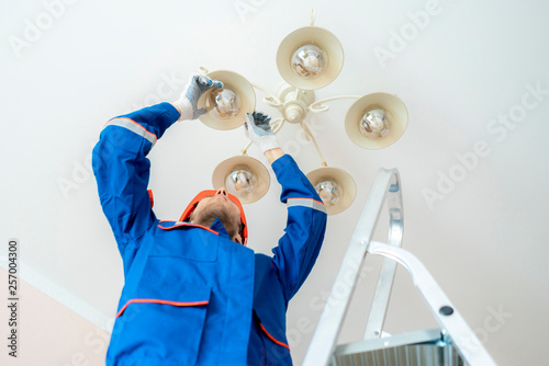 worker in uniform standing on the ladder and repair the chandelier at home f