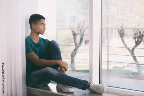 Upset African-American teenage boy sitting alone near window photo