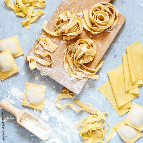 Homemade italian pasta, ravioli, fettuccine, tagliatelle on a wooden board and on a blue background. The cooking process, raw pasta. Tasty raw ravioli with ricotta and spinach,with flour on background