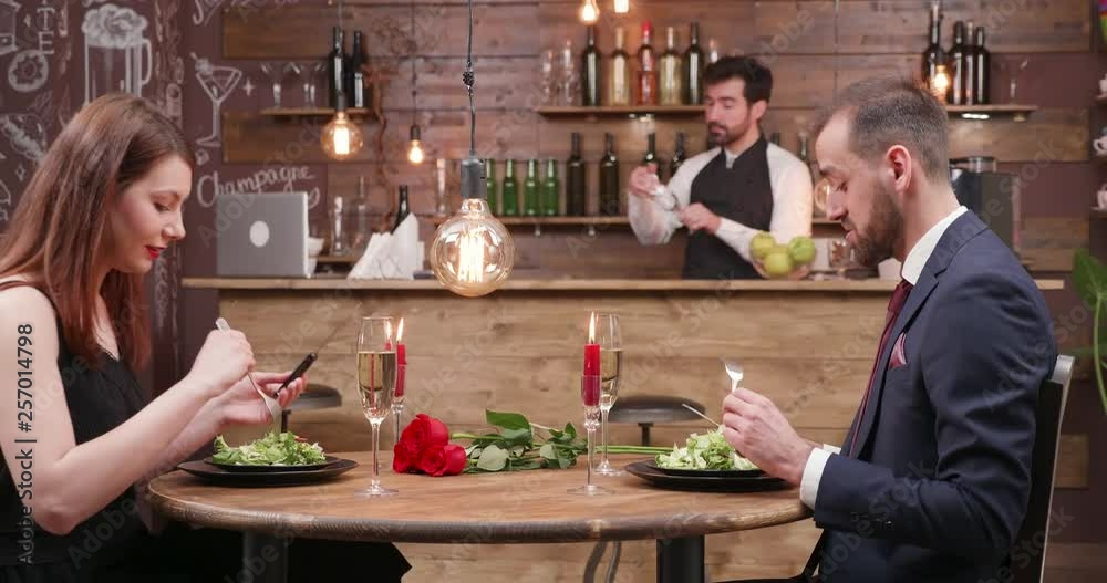 Young beautiful couple enjoying a night out. eating salad at a dinner table. Romantic candles and red roses on a wooden table.