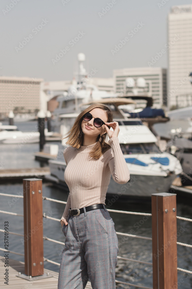Young european woman with blond long bob hair, beige turtleneck and trousers with pockets standing near yacht or boat in a sunny day