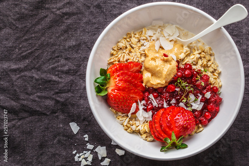 Oatmeal porridge with berries, peanut butter and coconut in white bowl, dark background. Healthy vegan food concept.