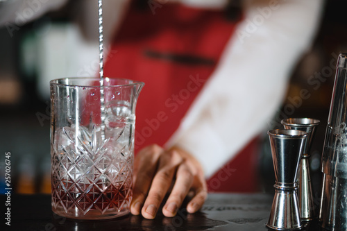 Barman mixing cocktail ingredients with a long spoon in high glass filled with ice cubes on the bar counter, close up