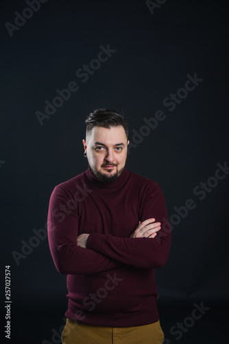 a man with a beard in casual clothes poses on an isolated dark background, in a burgundy sweater in the studio