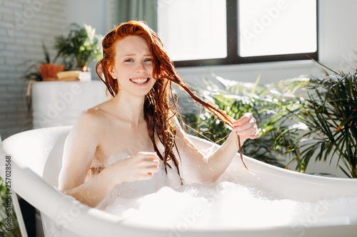 Playful young caucasian woman smiling, enjoying relaxing foam bath at home. Resting, relaxing, touhing her wet ginger hair, smiling. photo