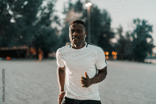 Smiling african male runner in white t-shirt running at the beach. Earphones in ears. One run can change your day  many runs can change your life.
