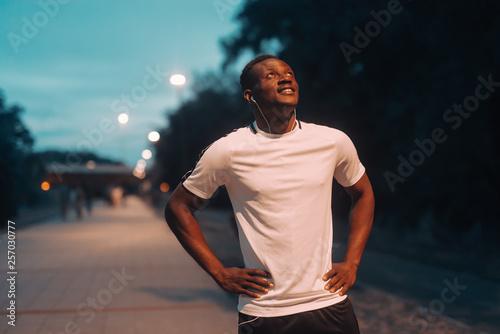 Smiling handsome african runner in sportswear holding hands on hips and looking up. Night workout concept. I will, just watch me.