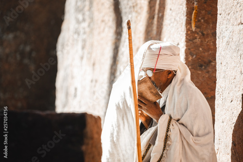 Old priest in white robe with walking stick and glasses reading the bible at the lalibela church in ethiopia 