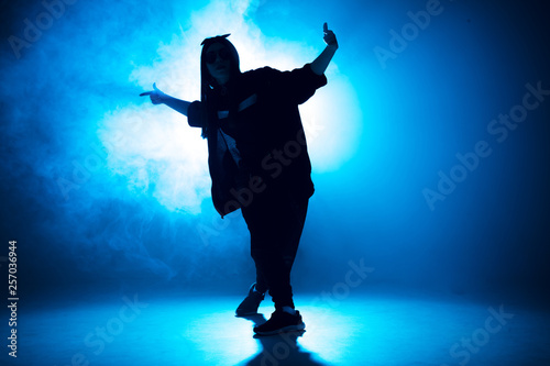Young woman silhouette dressed in street fashion wear dancing hip-hop style over studio blue light background with flare effects