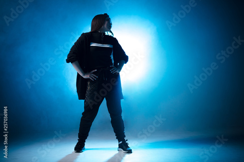 Young woman dressed in street fashion wear dancing hip-hop style over studio blue light background with flare effects