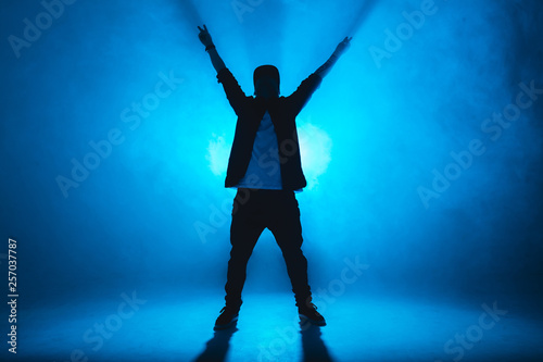 Stylish young man wearing trendy streetwear is dancing contemporary street dance on a blue studio background