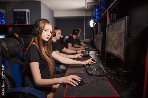 A cute female gamer girl with headset sits in a dark room behind a computer with a raw of another gamers sitting in line beside her in computer club. They are playing online games.