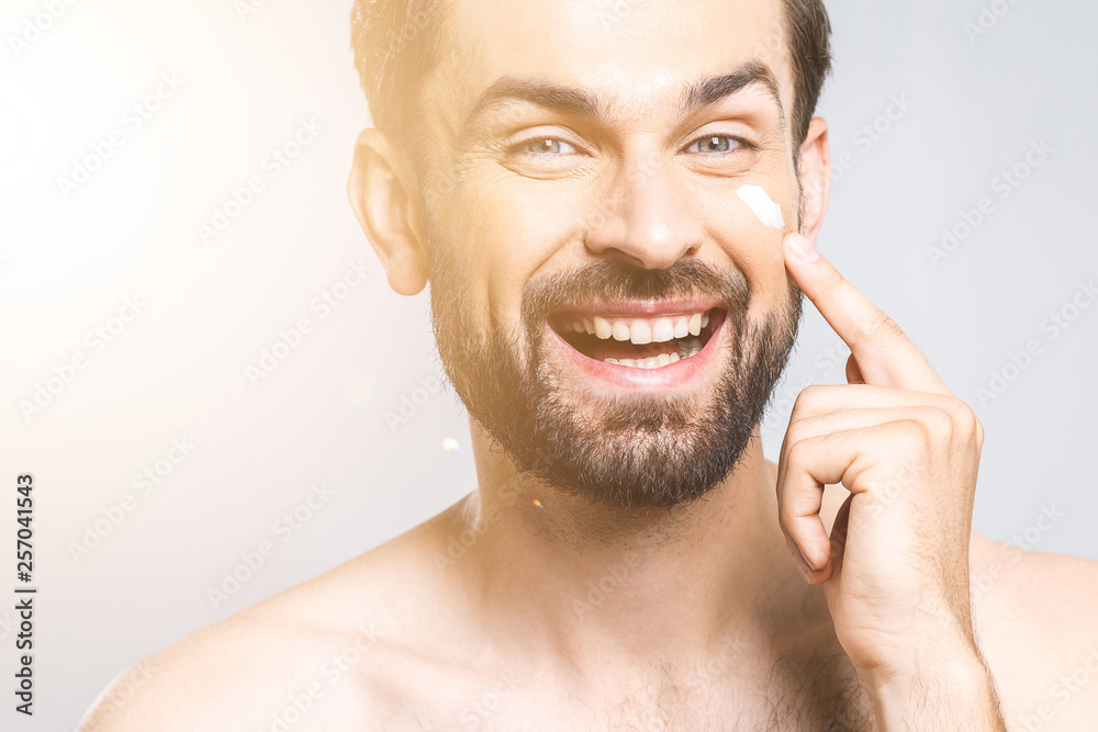 Obraz premium Skin care. Handsome happy young shirtless man applying cream at his face and looking at himself with smile while standing over gray background and looking at camera. Close-Up. Space for text.