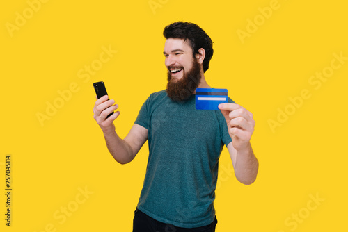 Young happy bearded man is buying through smart phone with his credit card on yellow background.