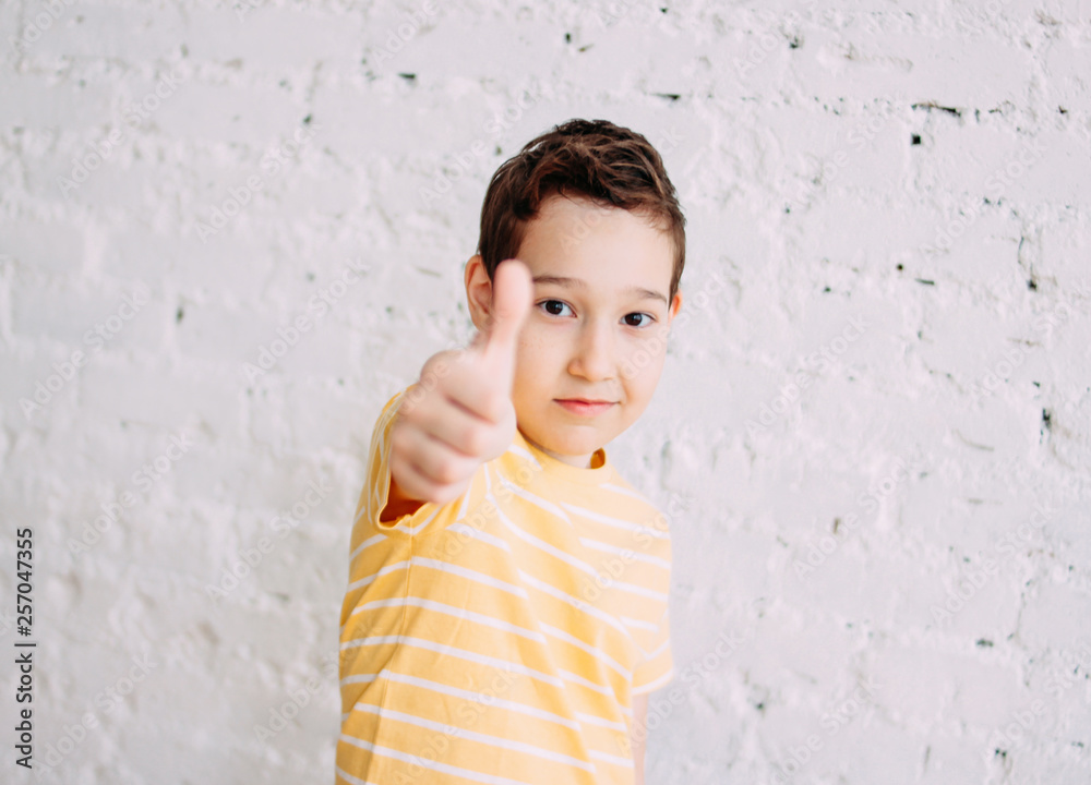 Portrait of cute tween smiling boy in yellow t-shirt shows thumb up isolated on white brick wall background Portrait of cute tween smiling boy in yellow t-shirt shows thumb up isolated on white brick wall background