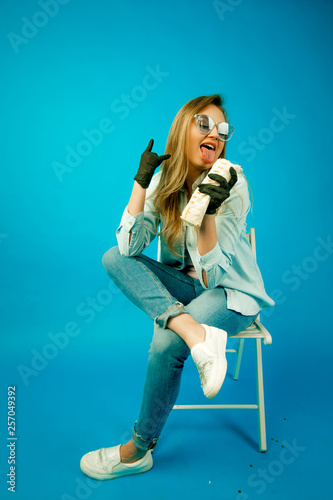 young beautiful blonde girl eating Fast food, a woman with an appetite eating Shawarma on a blue background, in denim clothes and glasses