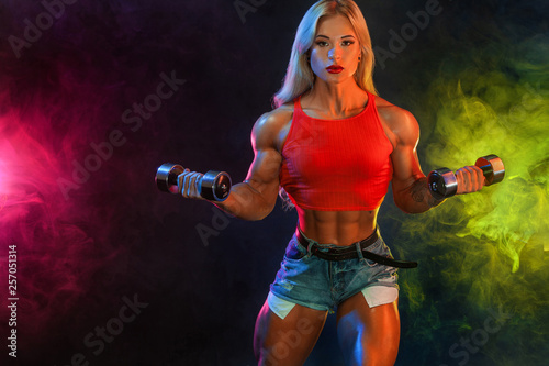 Strong athletic woman bodybuilder champion with dumbbells on dark background wearing in sportswear. Fitness and sport motivation.