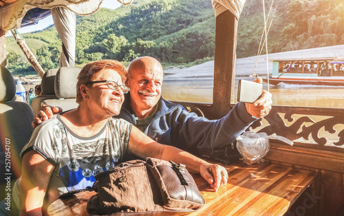 Senior retired couple of happy vacationers taking selfie at Mekong exploration tour with slowboat in Laos PDR - Active elderly travel concept on trip around the world - Warm saturated sunset filter photo