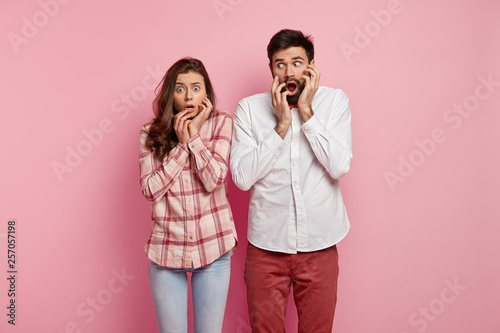 Image of scared speechless young girlfriend and boyfriend gasp with dropped jaws, concerned from surprise, stare at camera in amazement, stand next to each other, isolated over pink background