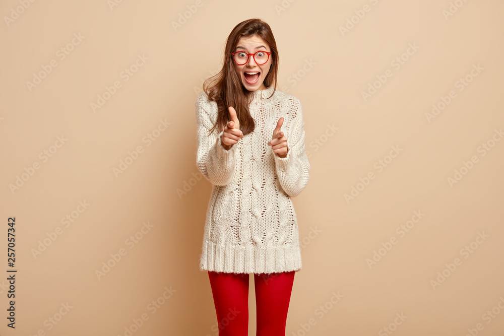 Horizontal view of joyful emotional woman makes finger gun geture, indicates at you, expresses nice choice, dressed in white jumper and red leggings, gestures happily, chooses you, stands indoor Horizontal view of joyful emotional woman makes finger gun geture, indicates at you, expresses nice choice, dressed in white jumper and red leggings, gestures happily, chooses you, stands indoor