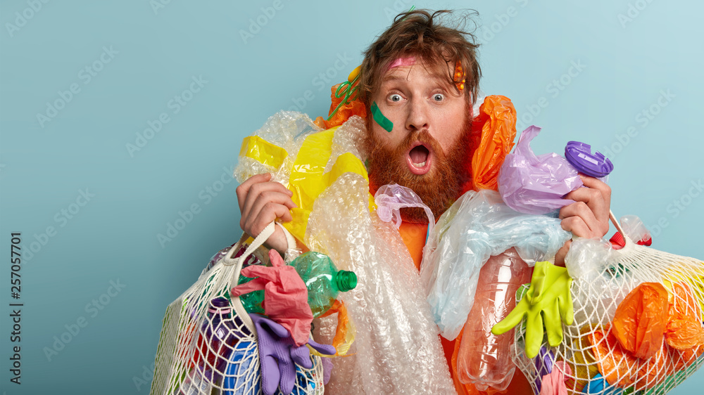 Emotive red haired man with thick bristle, carries much garbage, fights against plastic pollution, has shocked facial expression, isolated over blue background with blank space for your text