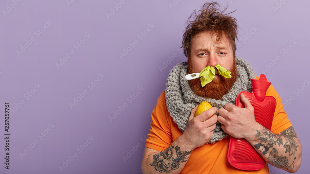Photo of sick man has terrible sneezing, keeps napkins in nose, suffers from rhinitis, holds lemon and hot water bottle, keeps thermometer in mouth, has seasonal influenza virus, isolated on lilac - obrazy, fototapety, plakaty 