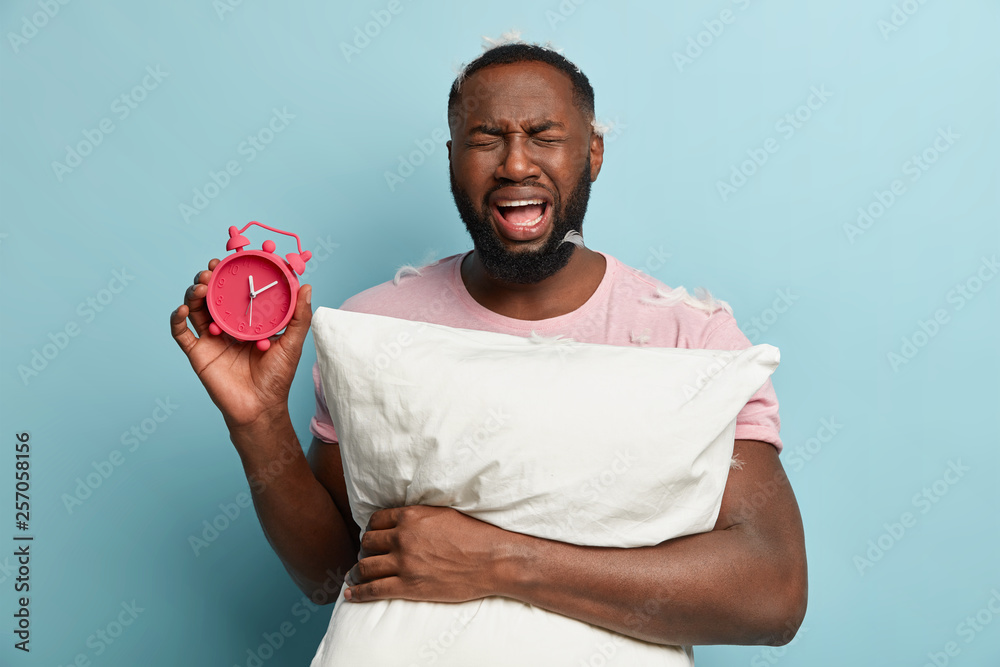 Depressed stressful black man with thick bristle, cries emotionally, wants to sleep well, carries red alarm clock and pillow, has sorrowful expressions, isolated on blue wall. Bad sleeping concept Depressed stressful black man with thick bristle, cries emotionally, wants to sleep well, carries red alarm clock and pillow, has sorrowful expressions, isolated on blue wall. Bad sleeping concept