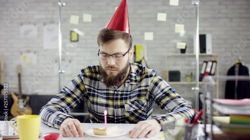 Sad attractive effective businessman celebrating a lonely birthday in the office, he is blowing a candle on a small cake. He is sitting in his office. Wearing formal suit, formal clothes. He is to eat photo