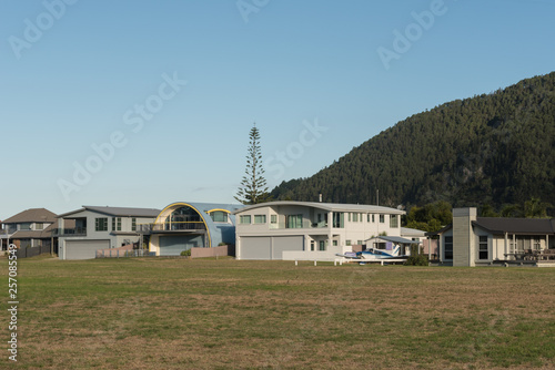 Fototapeta Naklejka Na Ścianę i Meble -  The grass airstrip at Pauanui flanked by residences with aircraft storage underneath, in late afternoon light. Pauanui, Coromandel Peninsula, New Zealand.